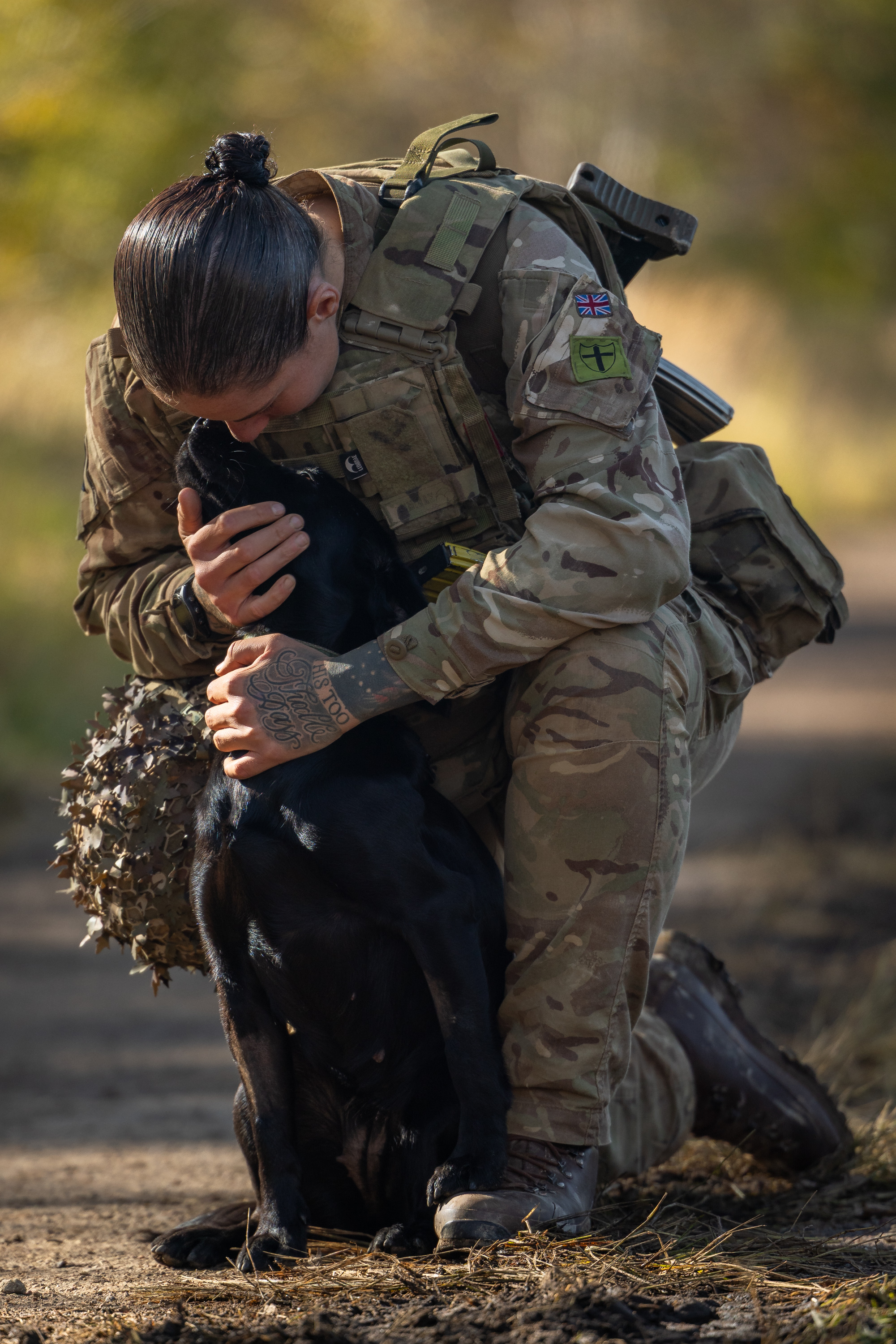 Soldier in camouflage uniform kneels and embraces a black dog affectionately outdoors on a dirt path.