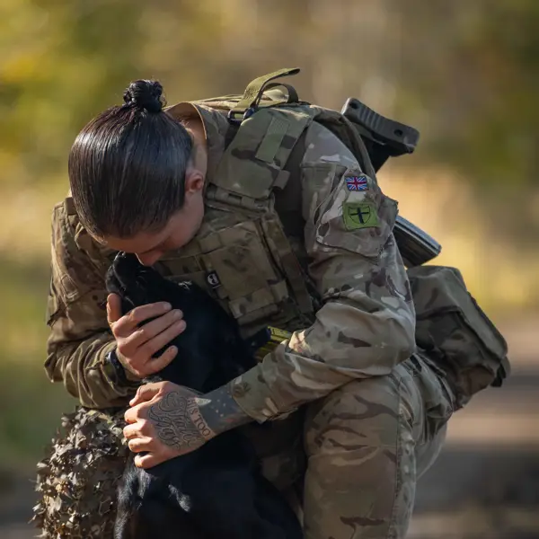 Soldier in camouflage uniform kneels and embraces a black dog affectionately outdoors on a dirt path.
