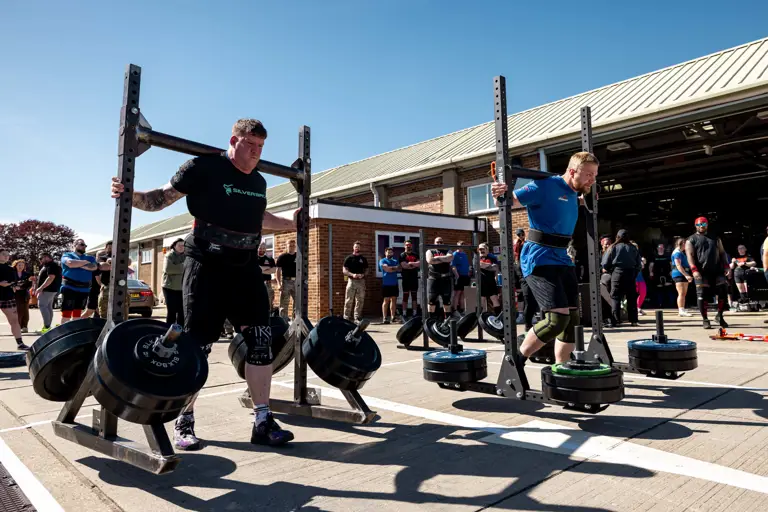 Two strongmen competing in a yoke carry event outdoors, each lifting heavy weights on metal frames. A crowd watches under a clear blue sky.