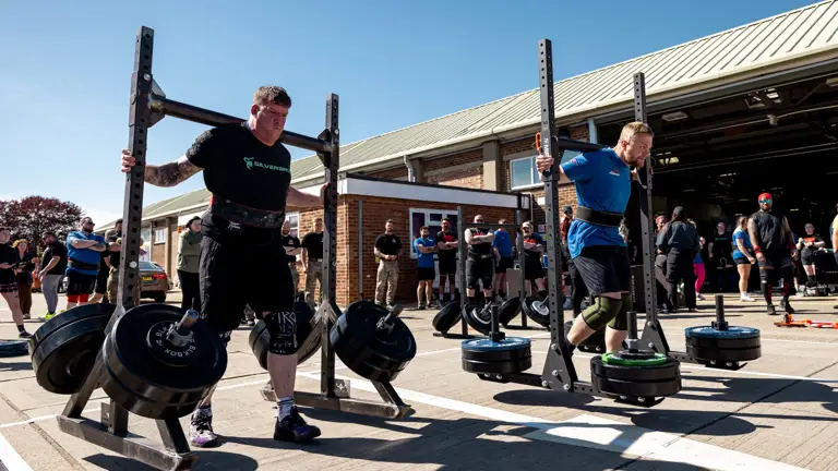 Two strongmen competing in a yoke carry event outdoors, each lifting heavy weights on metal frames. A crowd watches under a clear blue sky.