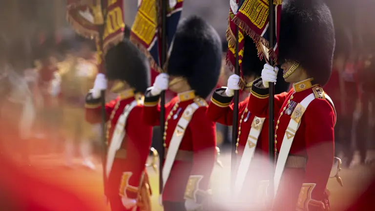 Four soldiers in red tunics with bearskin hats hold flags upright.