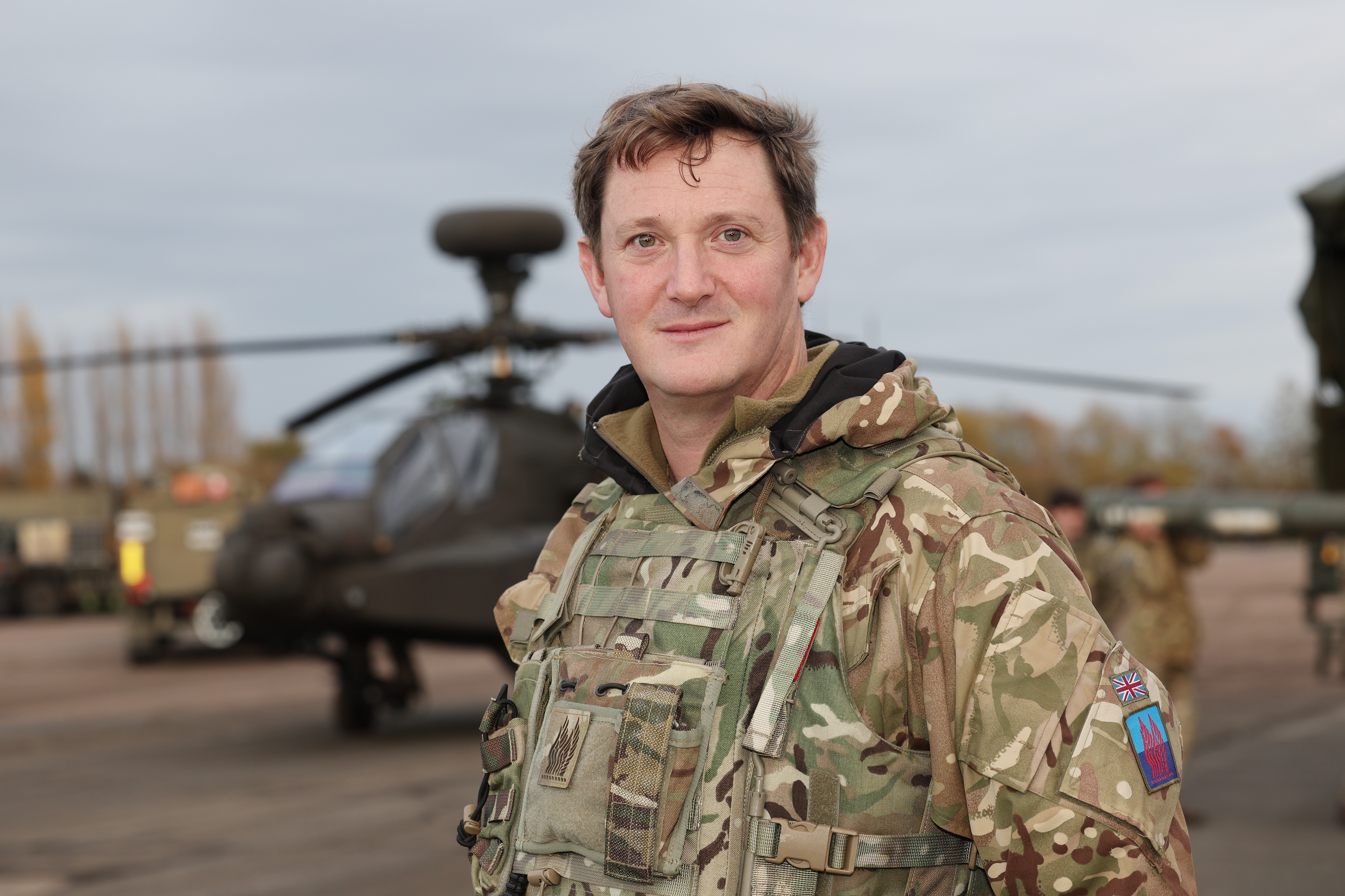 Officer in camouflage uniform stands on a tarmac with a military helicopter in the background during daytime.