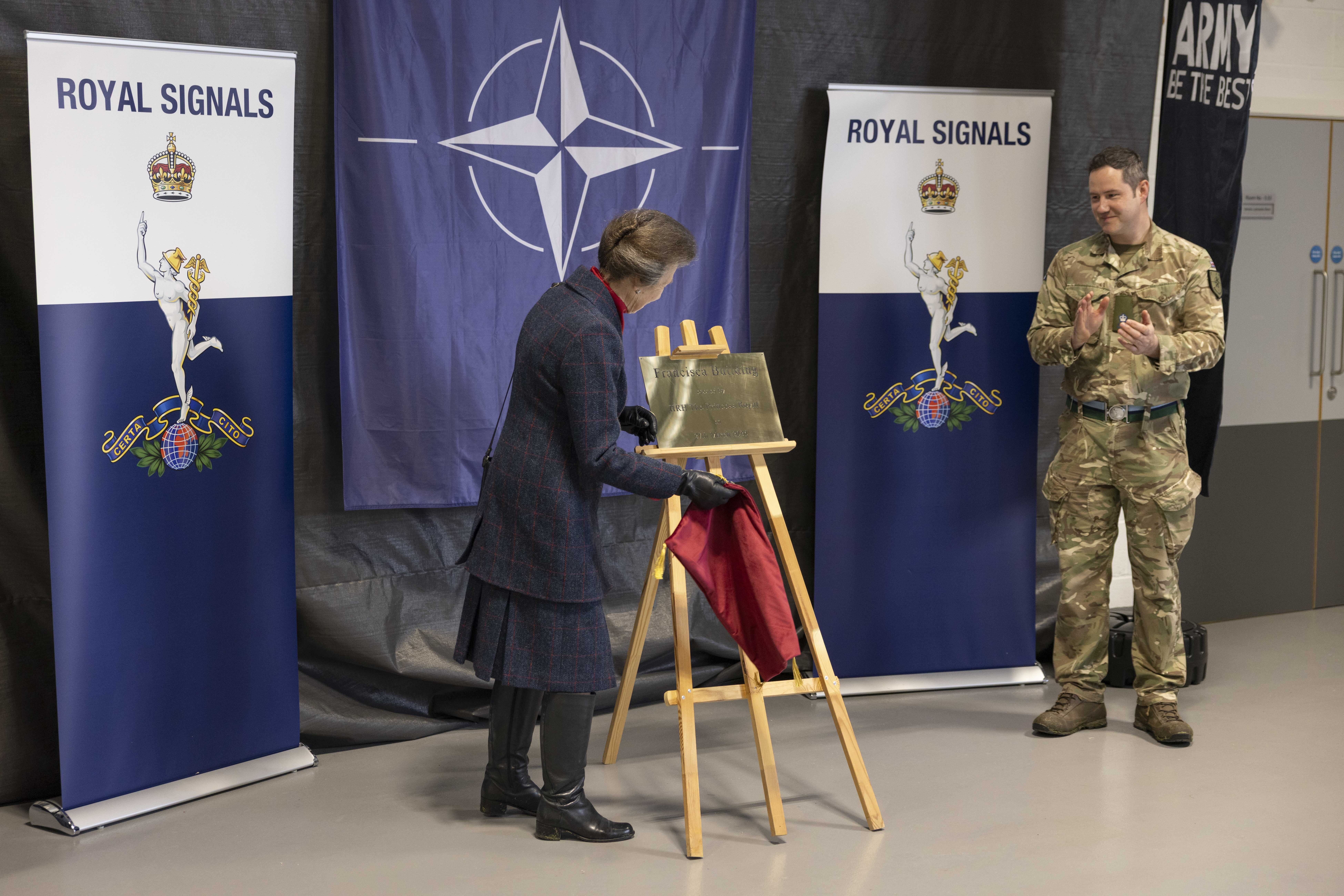 A person unveils a plaque on an easel, with NATO and Royal Signals banners in the background. Another person in military attire stands nearby, clapping.