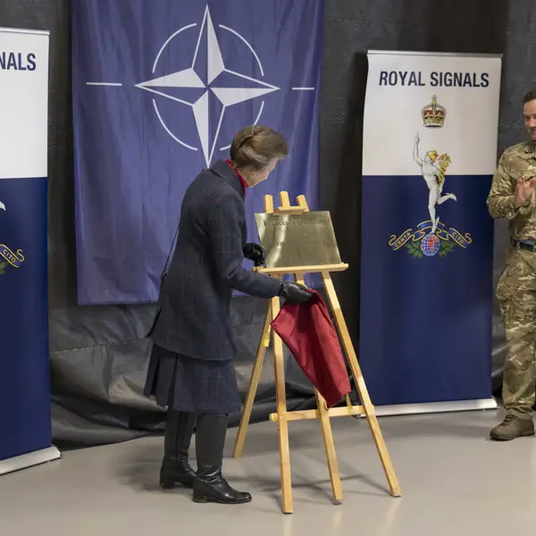 A person unveils a plaque on an easel, with NATO and Royal Signals banners in the background. Another person in military attire stands nearby, clapping.