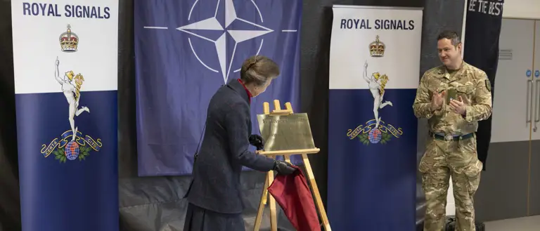 A person unveils a plaque on an easel, with NATO and Royal Signals banners in the background. Another person in military attire stands nearby, clapping.