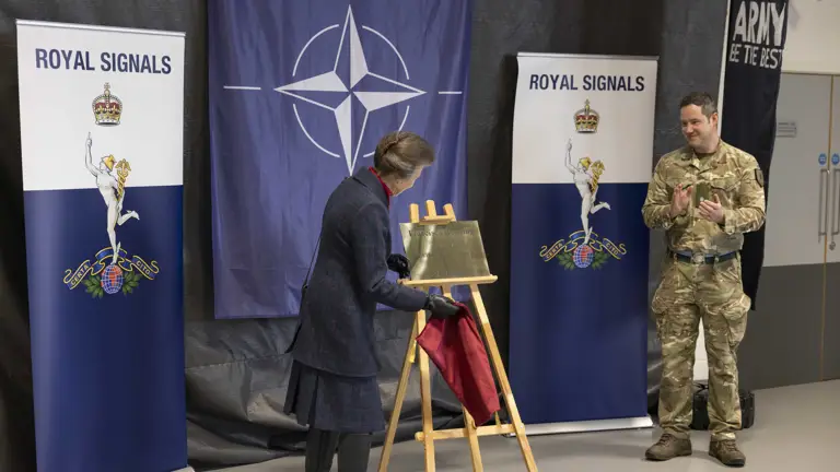 A person unveils a plaque on an easel, with NATO and Royal Signals banners in the background. Another person in military attire stands nearby, clapping.