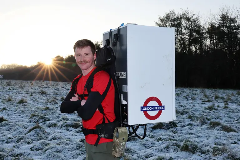 Major Joss Mowforth, pictured in his workout clothes posing for a portrait outside in snowy weather carrying a fridge strapped to his back. 