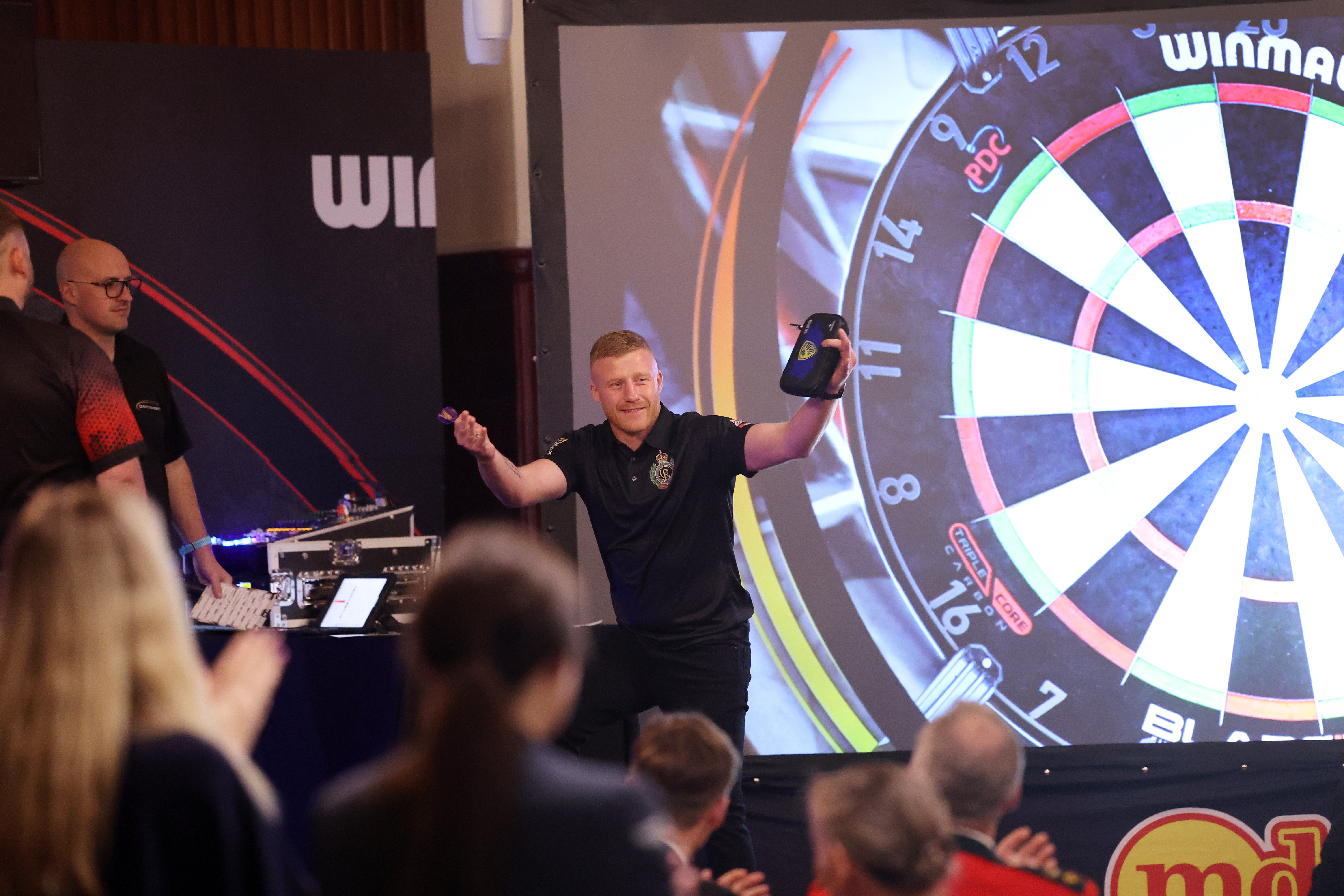 Player celebrating a successful dart throw in front of a large projected dartboard during a tournament.