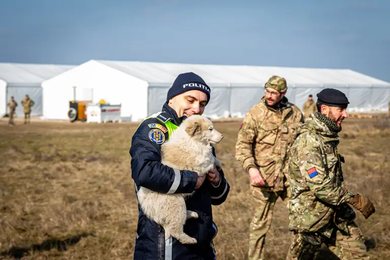 A Romanian police officer smiles at a puppy that he is holding, various military personnel can be seen in the background.