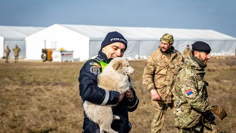 A Romanian police officer smiles at a puppy that he is holding, various military personnel can be seen in the background.