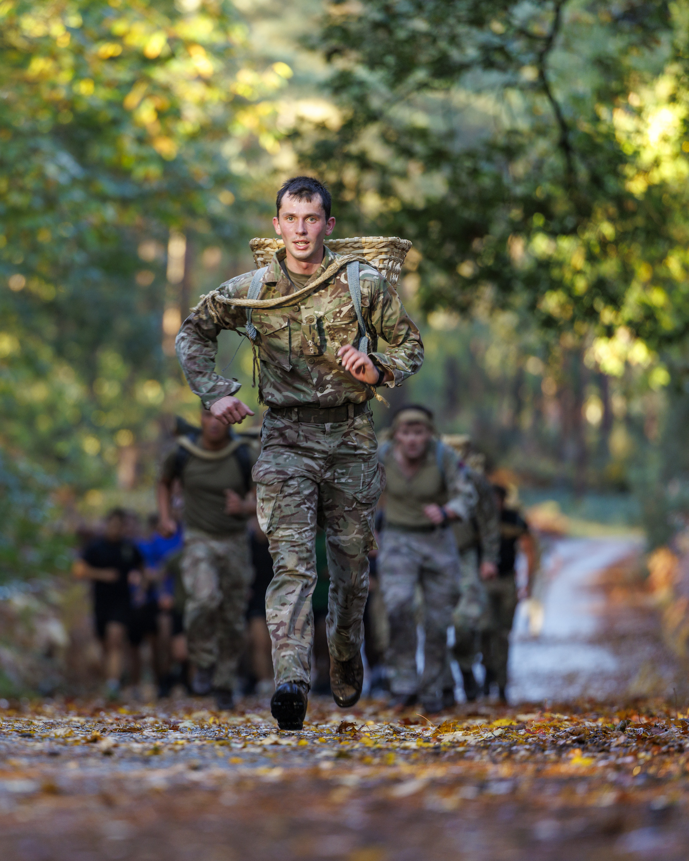 Soldier in camouflage uniform running on a forest trail carrying a large woven basket on his back during training.