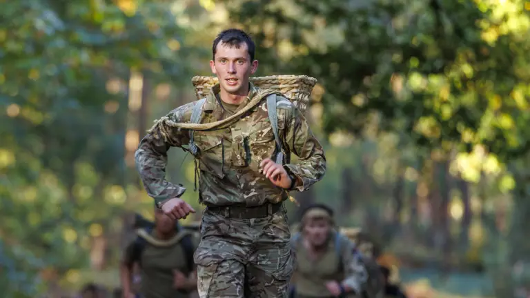 Soldier in camouflage uniform running on a forest trail carrying a large woven basket on his back during training.