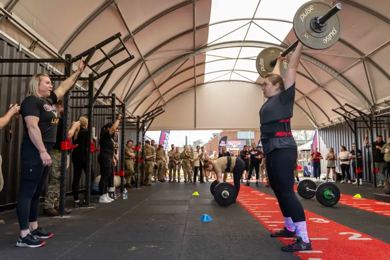 A person lifts a barbell overhead in a gym, surrounded by spectators and military personnel. The atmosphere is focused and supportive.