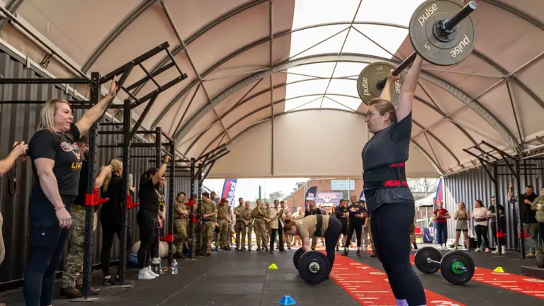 A person lifts a barbell overhead in a gym, surrounded by spectators and military personnel. The atmosphere is focused and supportive.