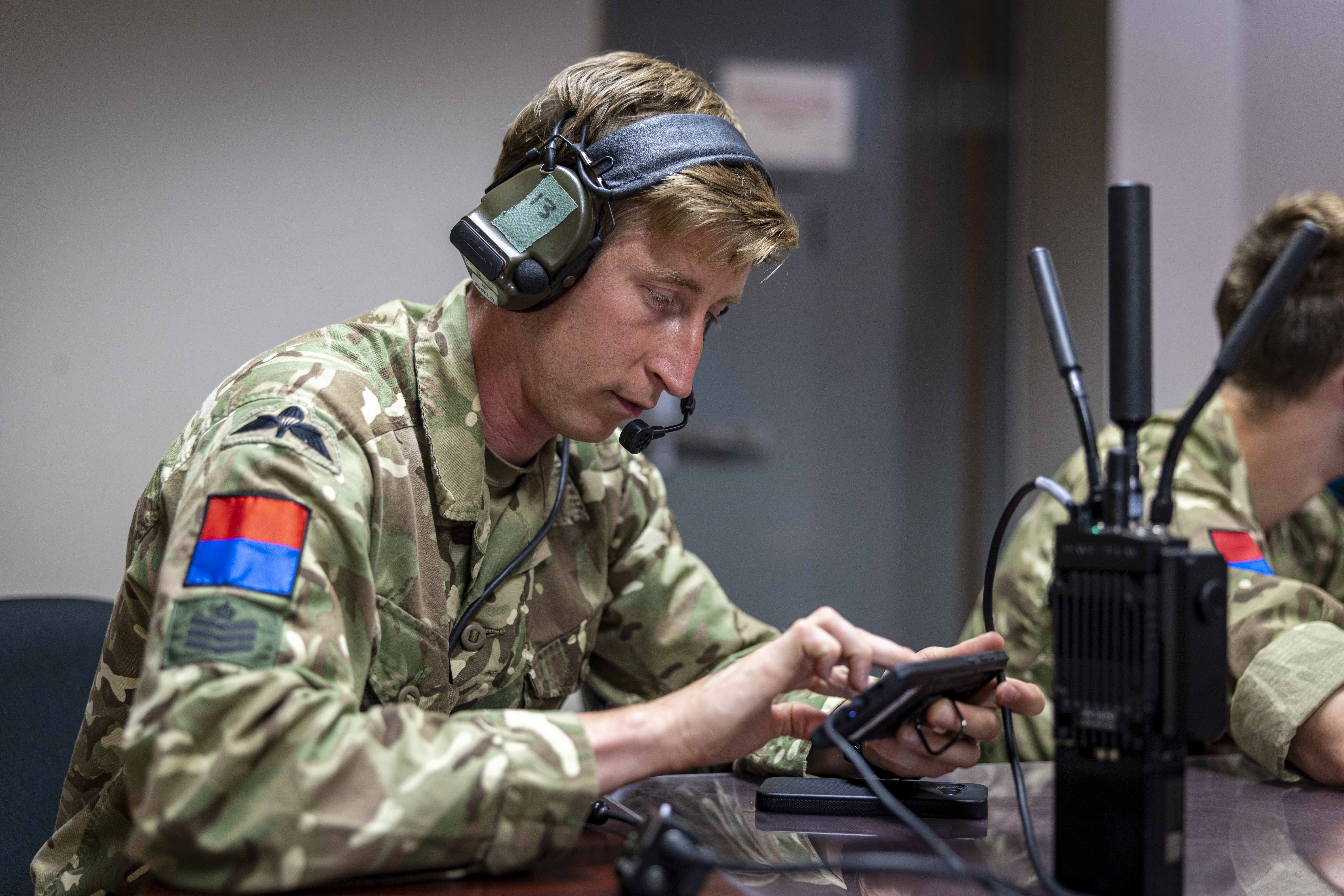 Military personnel in camouflage uniform using a portable communication device at a desk. The uniform has patches, including a flag and insignia. A radio with antennas is also on the desk.