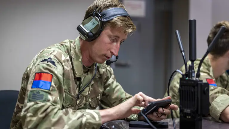 Military personnel in camouflage uniform using a portable communication device at a desk. The uniform has patches, including a flag and insignia. A radio with antennas is also on the desk.