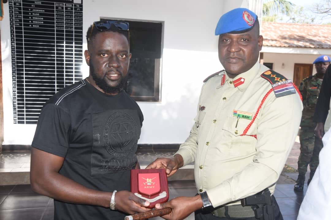 A military officer in uniform presents a red plaque with crossed swords and 'ARMY' inscription to a man in a black t-shirt.