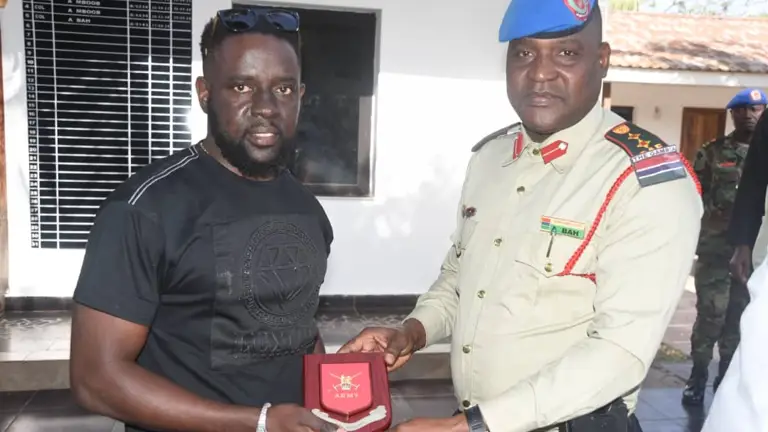 A military officer in uniform presents a red plaque with crossed swords and 'ARMY' inscription to a man in a black t-shirt.