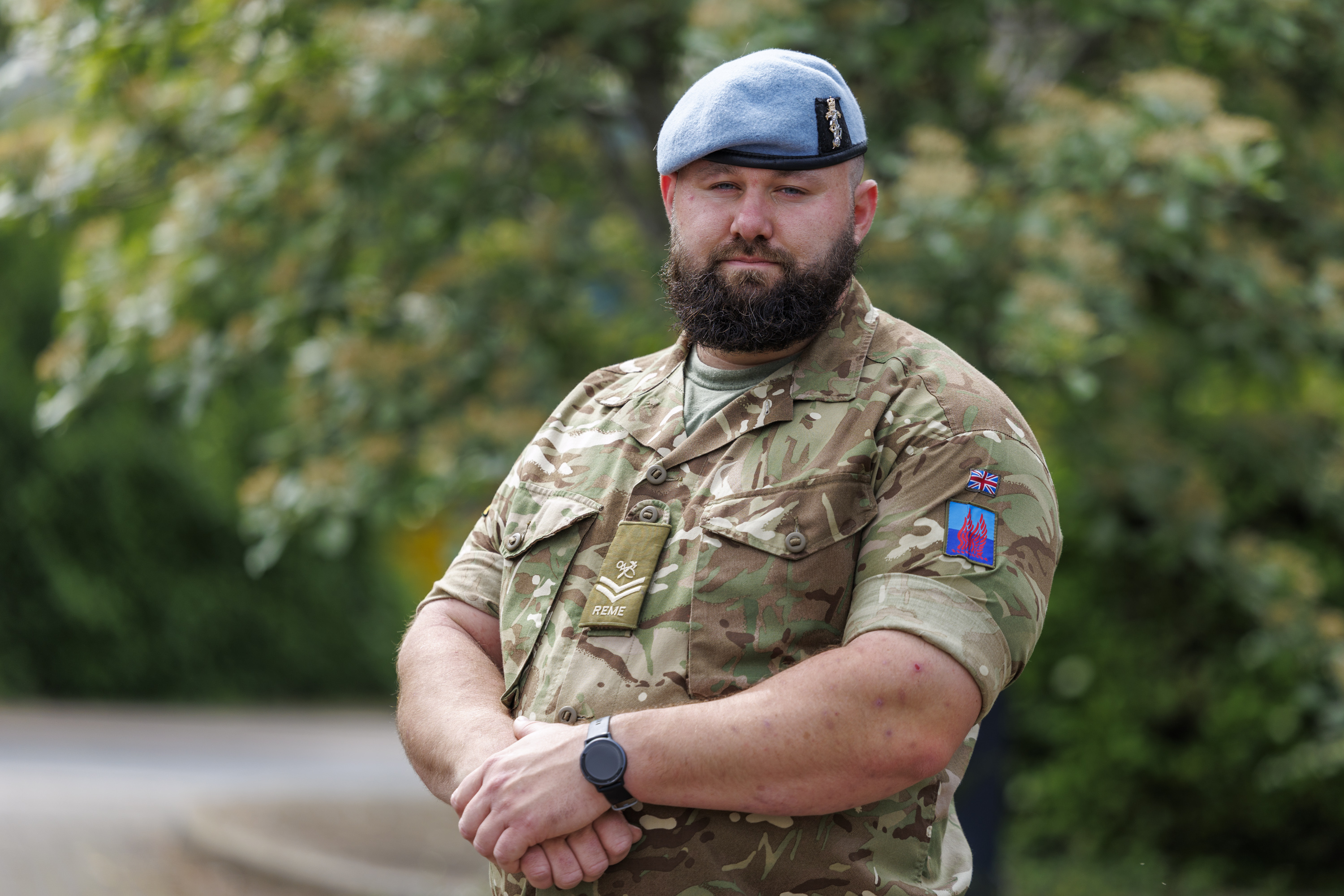 A person in a military camouflage uniform stands outdoors with arms crossed. They are wearing a blue beret and have a watch on one wrist. The background features leafy trees.