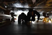 Two people crawling through a narrow, dimly lit tunnel with rough, rocky walls overhead.