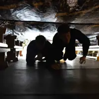 Two people crawling through a narrow, dimly lit tunnel with rough, rocky walls overhead.