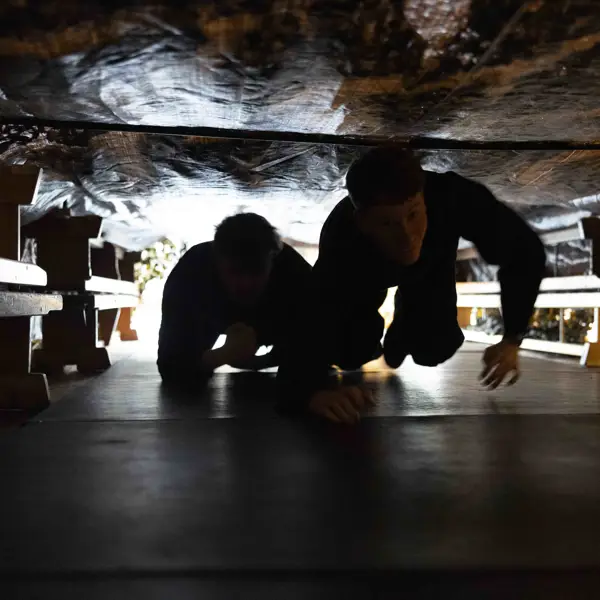 Two people crawling through a narrow, dimly lit tunnel with rough, rocky walls overhead.