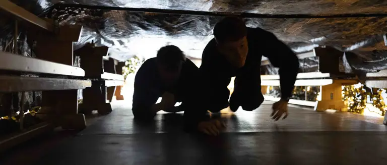 Two people crawling through a narrow, dimly lit tunnel with rough, rocky walls overhead.