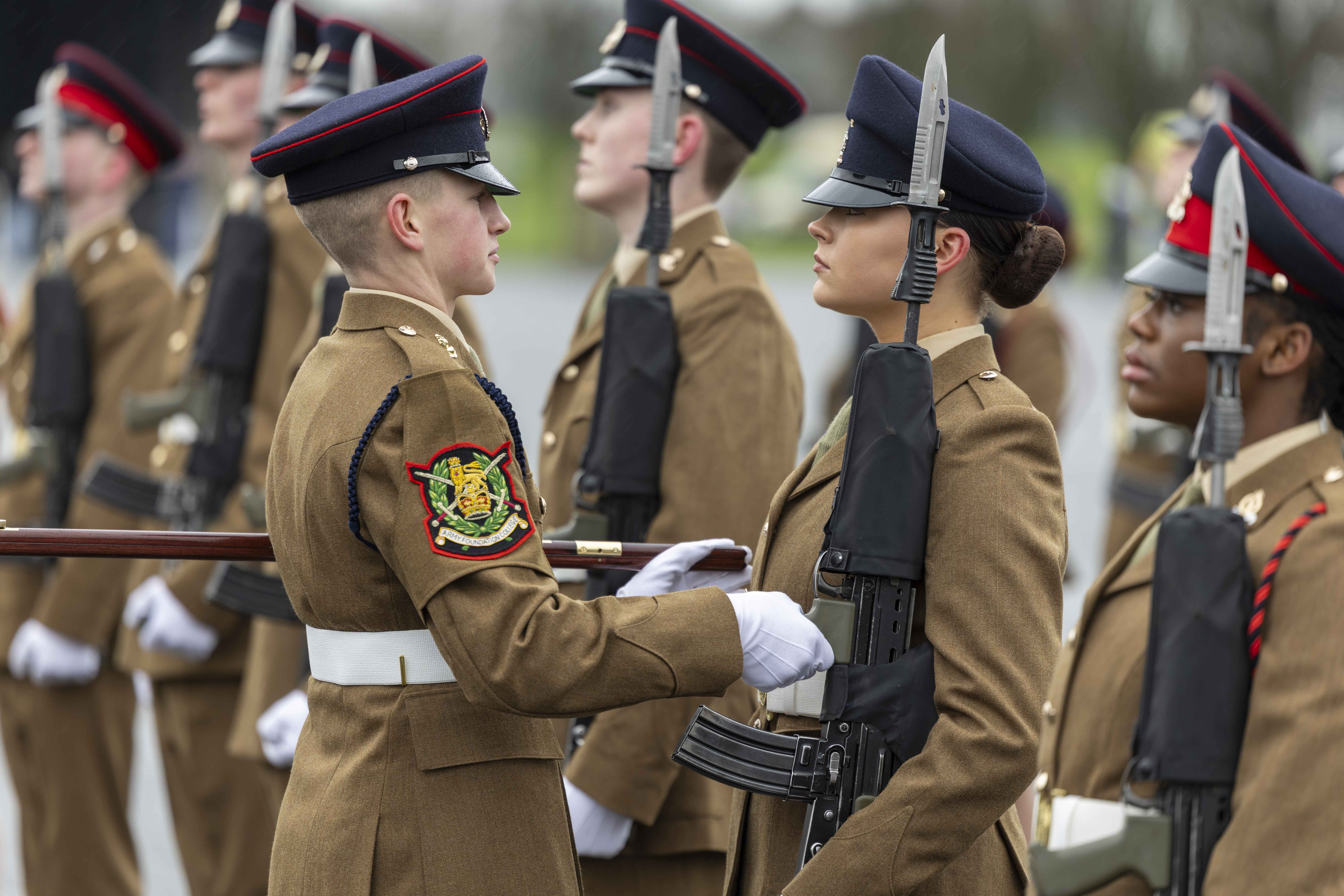 Soldiers in brown uniforms and peaked caps stand in formation, holding rifles with bayonets. One soldier inspects another, showing discipline and focus.