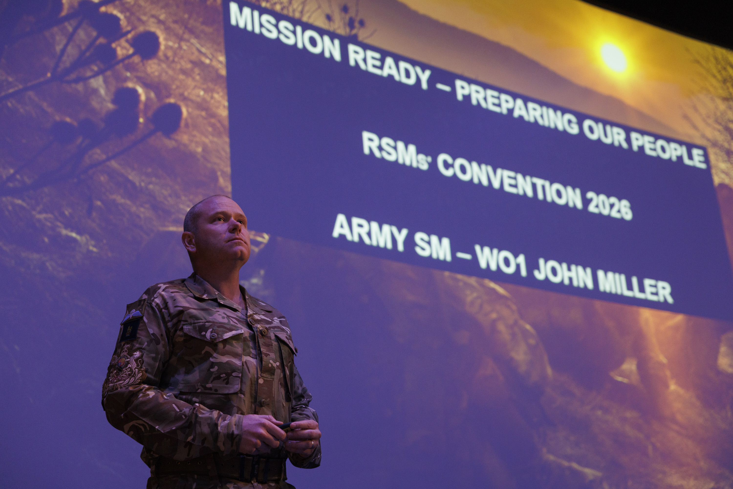 A person in military uniform stands confidently on stage in front of a large screen displaying text: 