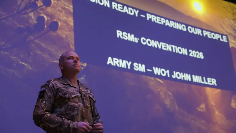 A person in military uniform stands confidently on stage in front of a large screen displaying text: