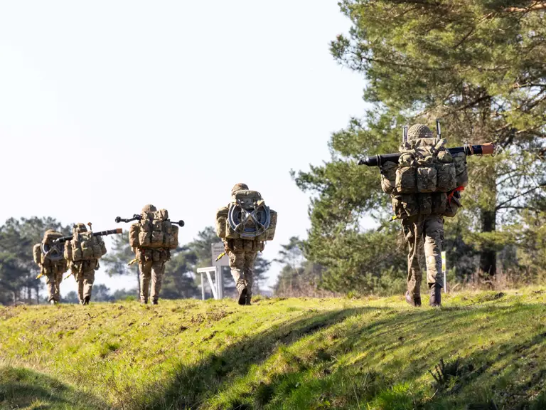 Soldiers in camouflage gear prepare equipment in a dry field while multiple parachutes descend from the clear blue sky.