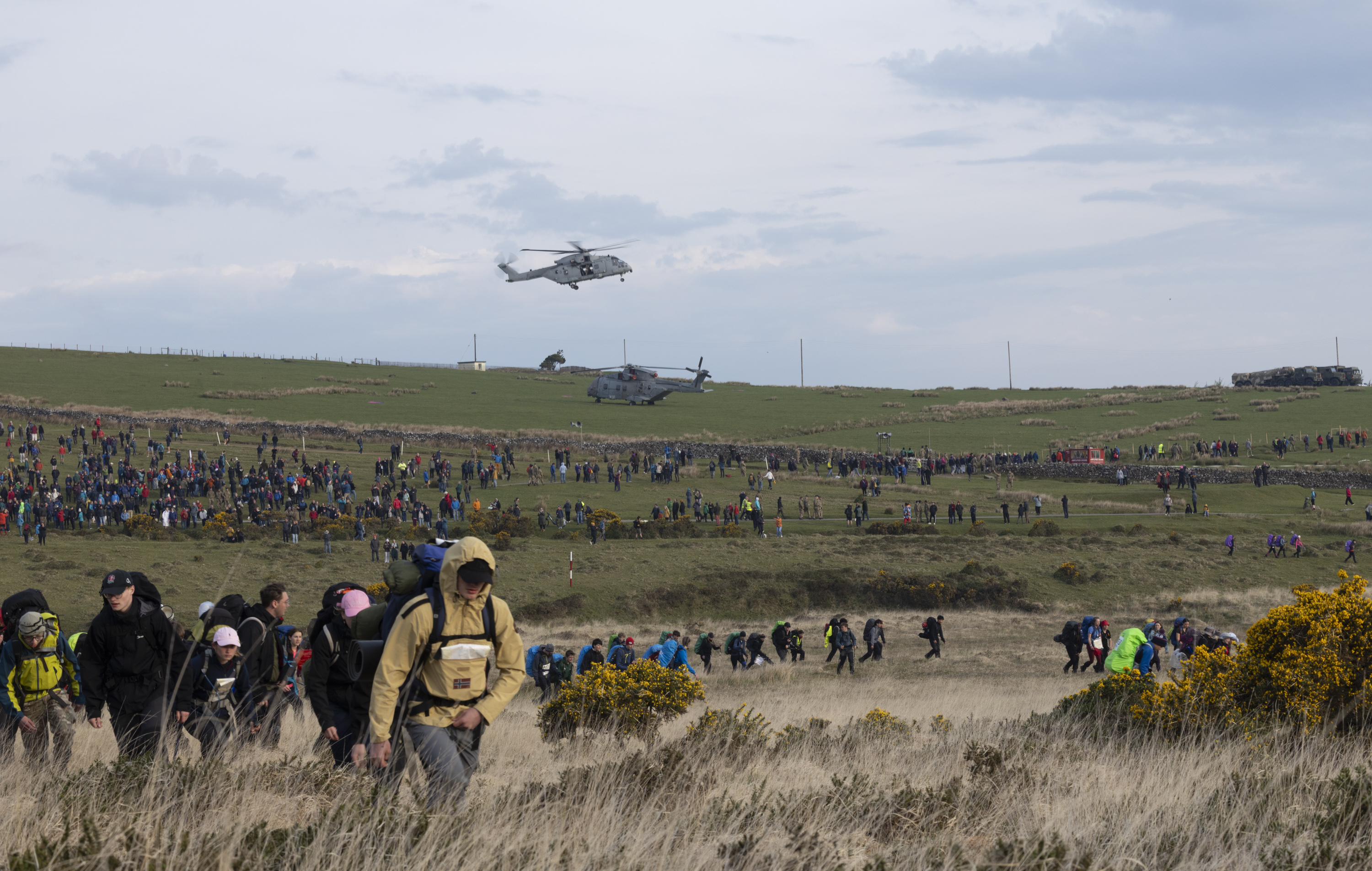 A large crowd of people hike across a grassy field under a cloudy sky, with two helicopters hovering above. The scene is bustling and energetic.