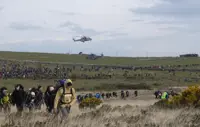 A large crowd of people hike across a grassy field under a cloudy sky, with two helicopters hovering above. The scene is bustling and energetic.