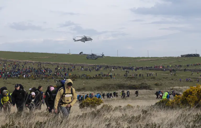 A large crowd of people hike across a grassy field under a cloudy sky, with two helicopters hovering above. The scene is bustling and energetic.