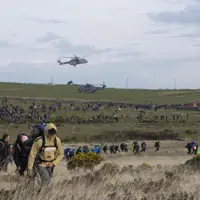 A large crowd of people hike across a grassy field under a cloudy sky, with two helicopters hovering above. The scene is bustling and energetic.