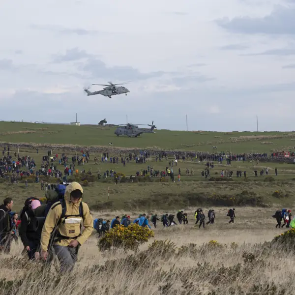 A large crowd of people hike across a grassy field under a cloudy sky, with two helicopters hovering above. The scene is bustling and energetic.