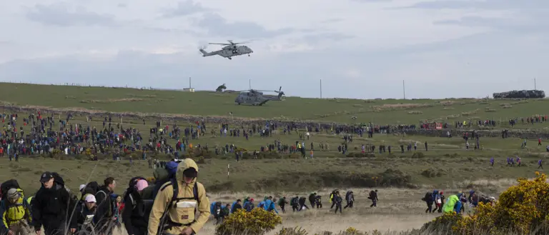 A large crowd of people hike across a grassy field under a cloudy sky, with two helicopters hovering above. The scene is bustling and energetic.