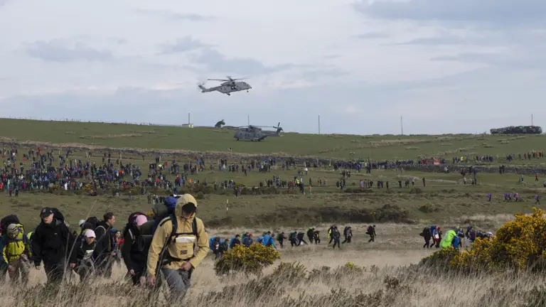 A large crowd of people hike across a grassy field under a cloudy sky, with two helicopters hovering above. The scene is bustling and energetic.