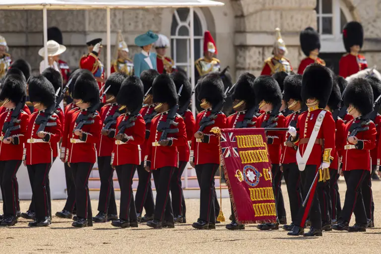 Two lines of soldiers in red tunics and bearskin hats stand with their rifles pointing upwards.