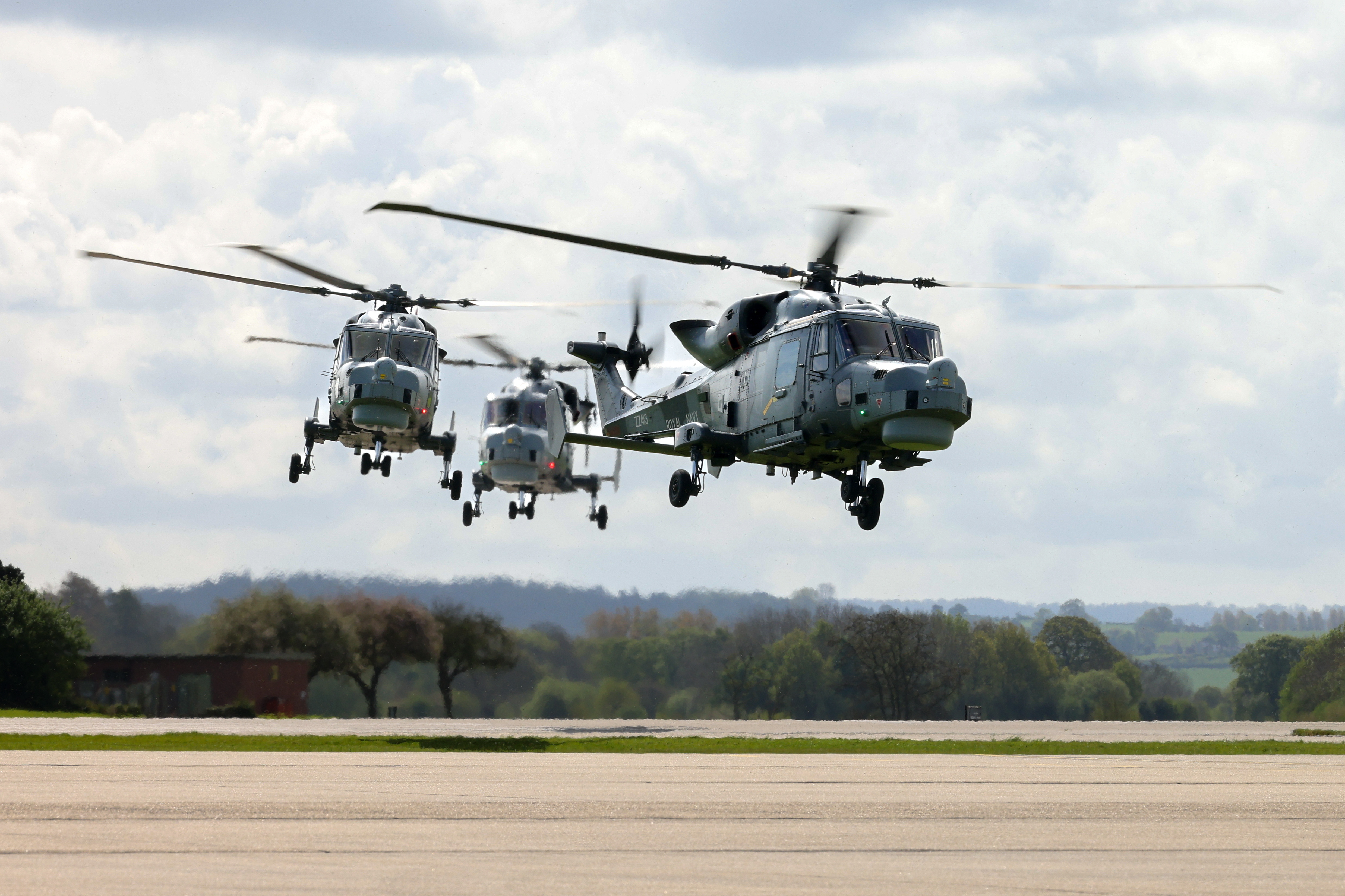 Three military helicopters are flying in formation above a runway. The sky is cloudy.