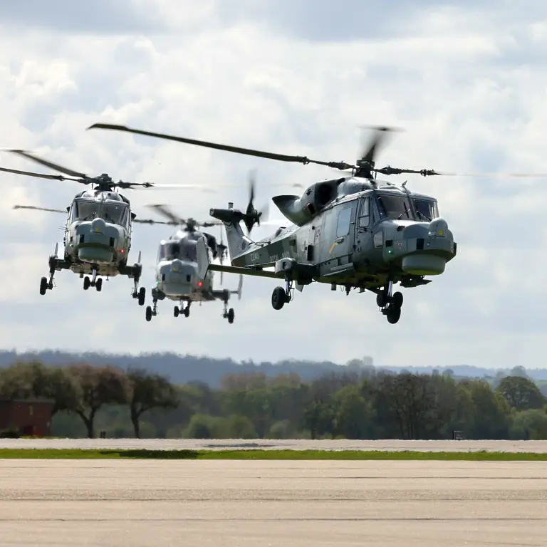 Three military helicopters are flying in formation above a runway. The sky is cloudy.