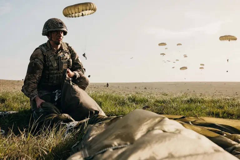 A soldier in camouflage uniform and helmet crouches on a grassy field, packing a parachute.
