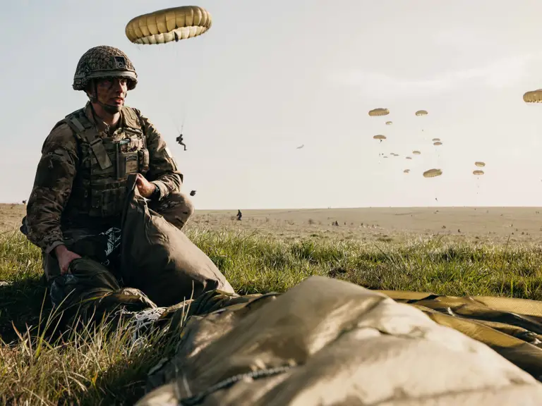 A soldier in camouflage uniform and helmet crouches on a grassy field, packing a parachute.