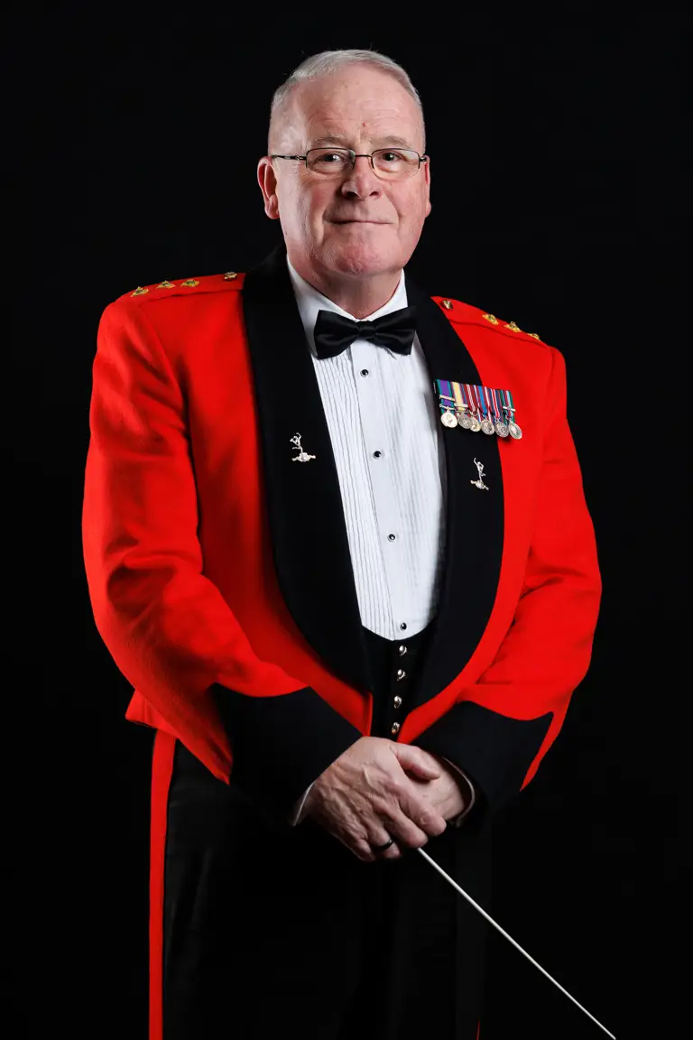 Man in a formal red military dress jacket with medals and a black bow tie holding a conductor's baton.
