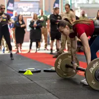 A woman in a red shirt prepares to lift a barbell in a gym competition. Onlookers, some in military attire, watch intently, creating an energetic atmosphere.