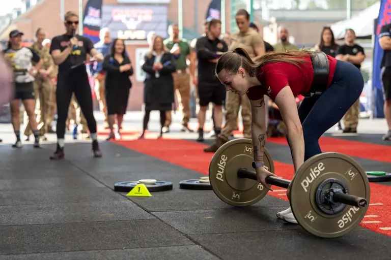 A woman in a red shirt prepares to lift a barbell in a gym competition. Onlookers, some in military attire, watch intently, creating an energetic atmosphere.