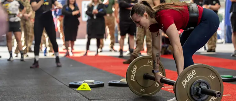 A woman in a red shirt prepares to lift a barbell in a gym competition. Onlookers, some in military attire, watch intently, creating an energetic atmosphere.
