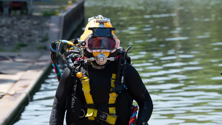 A diver in full gear with yellow helmet and breathing apparatus emerging from water near dock.