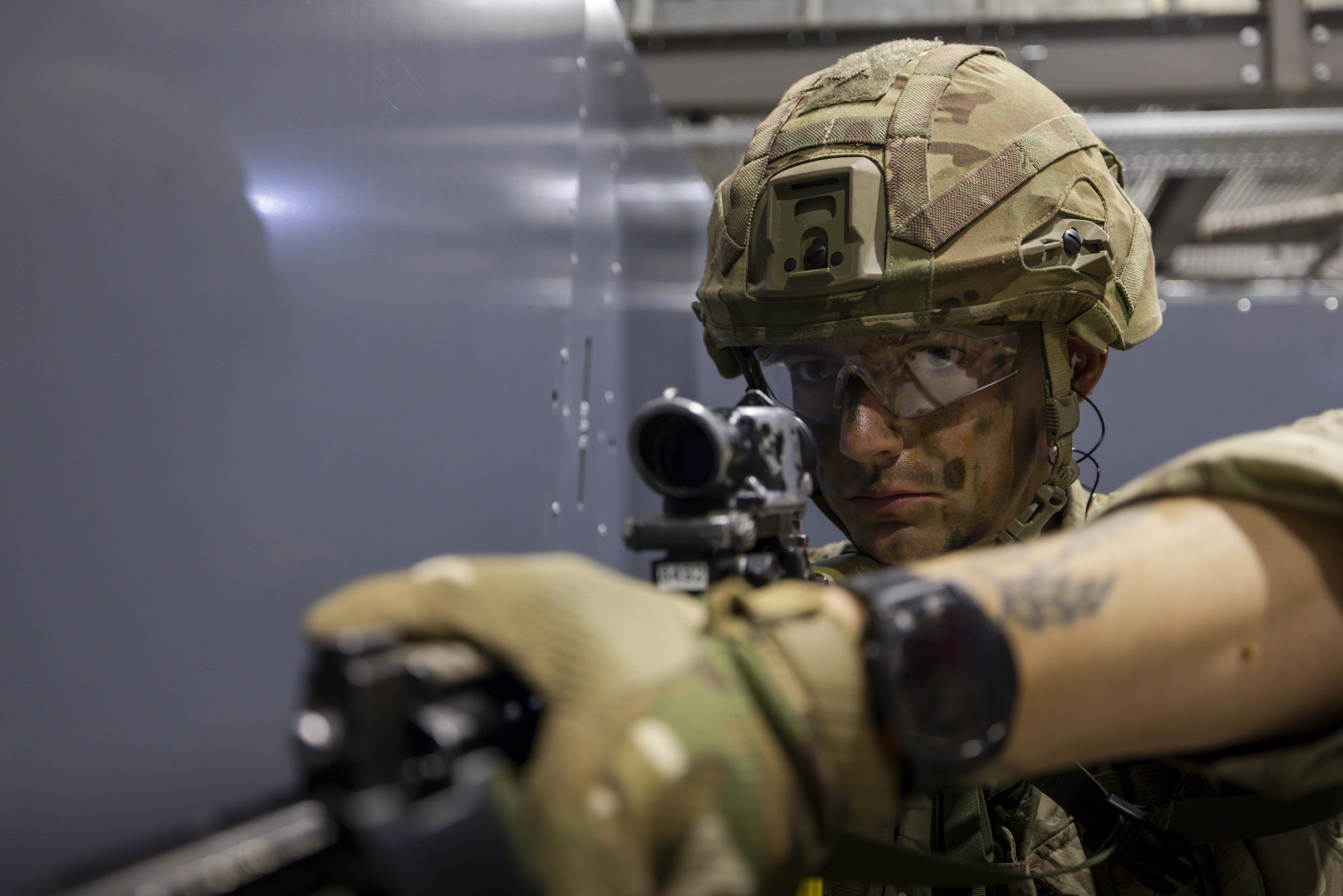 Military personnel in camouflage uniforms participate in a training exercise. One person holds a firearm and faces another individual, who is also armed. The environment appears to be indoors with dim lighting.