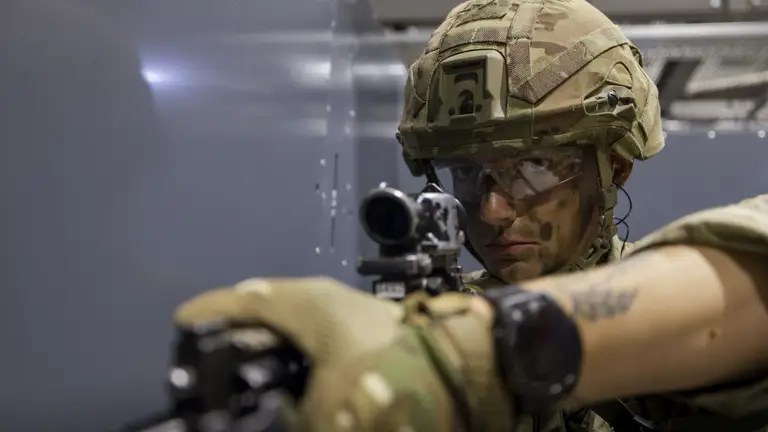 Military personnel in camouflage uniforms participate in a training exercise. One person holds a firearm and faces another individual, who is also armed. The environment appears to be indoors with dim lighting.
